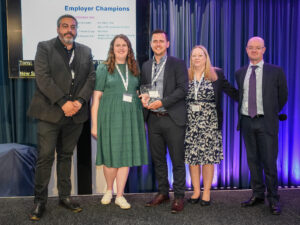 A photo of 5 people on stage being presented with an award. They are all facing the camera and smiling.