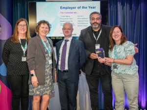 A photo of 5 people on stage. 1 person is presenting a glass award to another. Everyone is facing the camera and smiling.