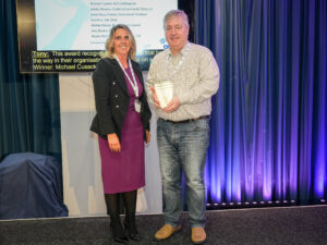 A photo of 2 people on stage. 1 person is holding a glass award and both are facing the camera and smiling. 