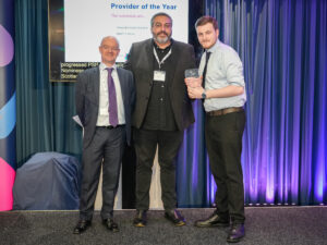 A photo of 3 people on stage. The person on the right is holding a glass award. They are all facing the camera and smiling.