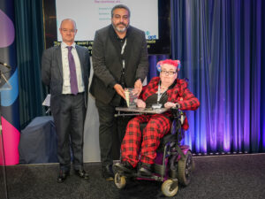 A photo of 3 people on stage. The person in the middle is presenting a glass award to person on the right They are all looking at the camera and smiling.