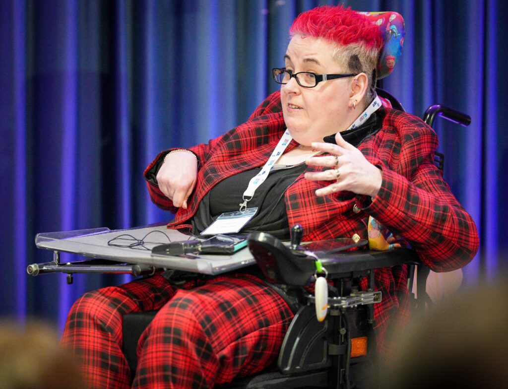 A photo of a woman who uses a wheelchair and is wearing a red tartan suit while presenting in front of an audience.
