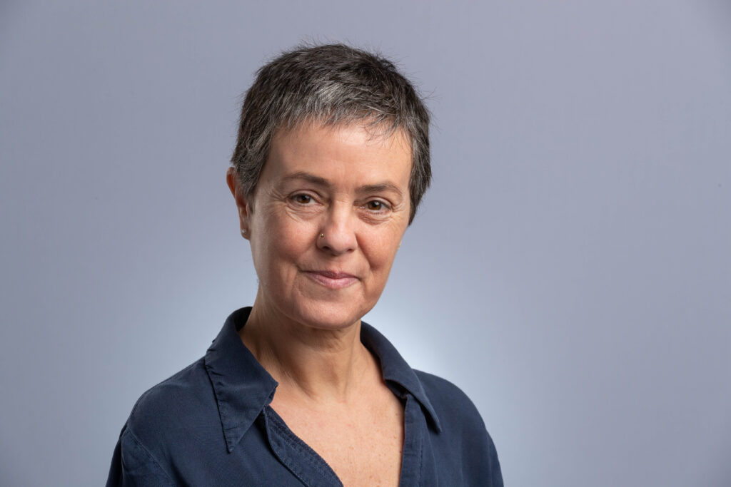 Head and shoulders image of a person with short hair and a dark shirt in front of a grey background.