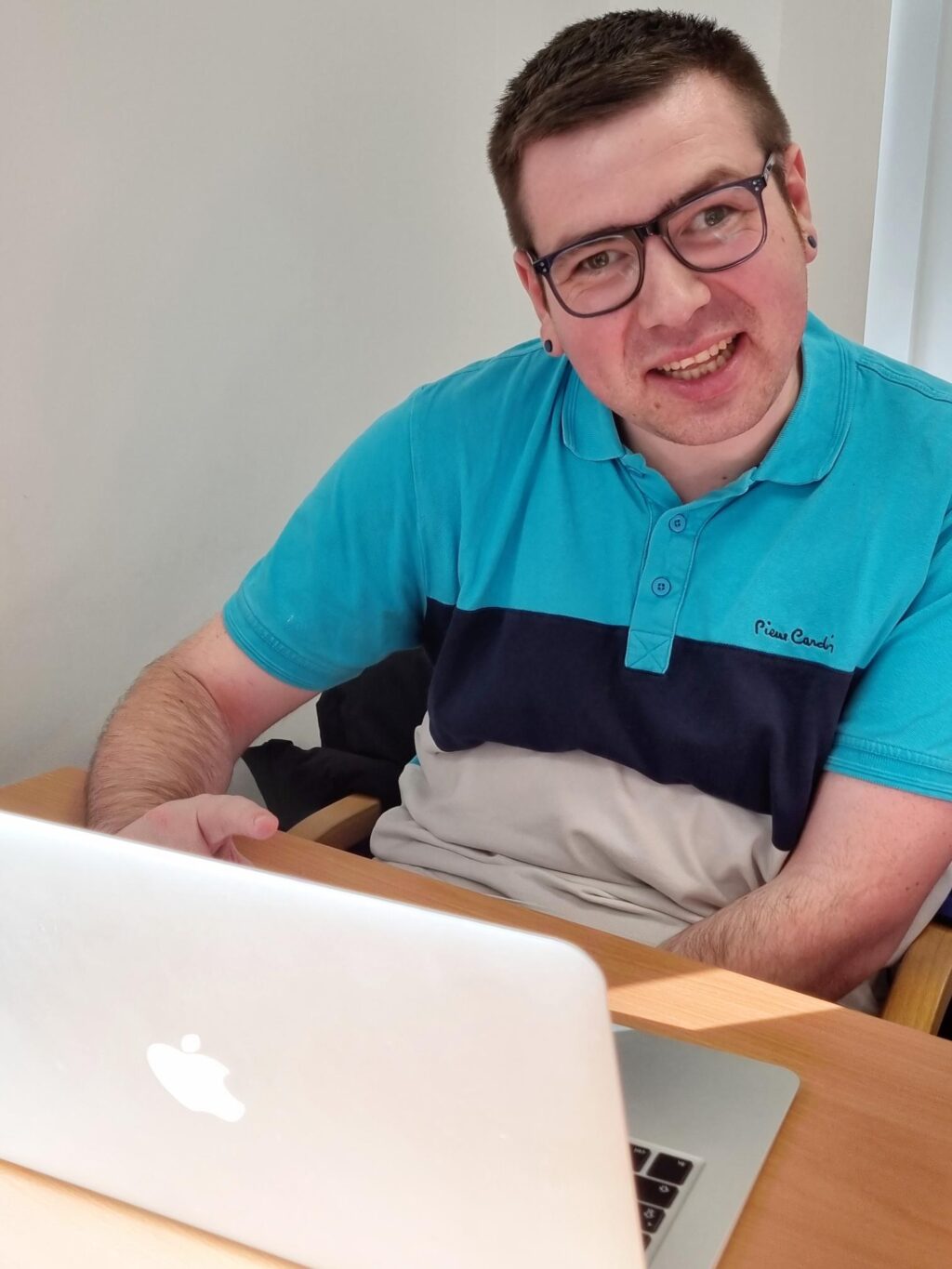 Kris McLaughlin has short dark hair and wears glasses. He is wearing a blue and white polo shirt and sitting in front of a laptop.